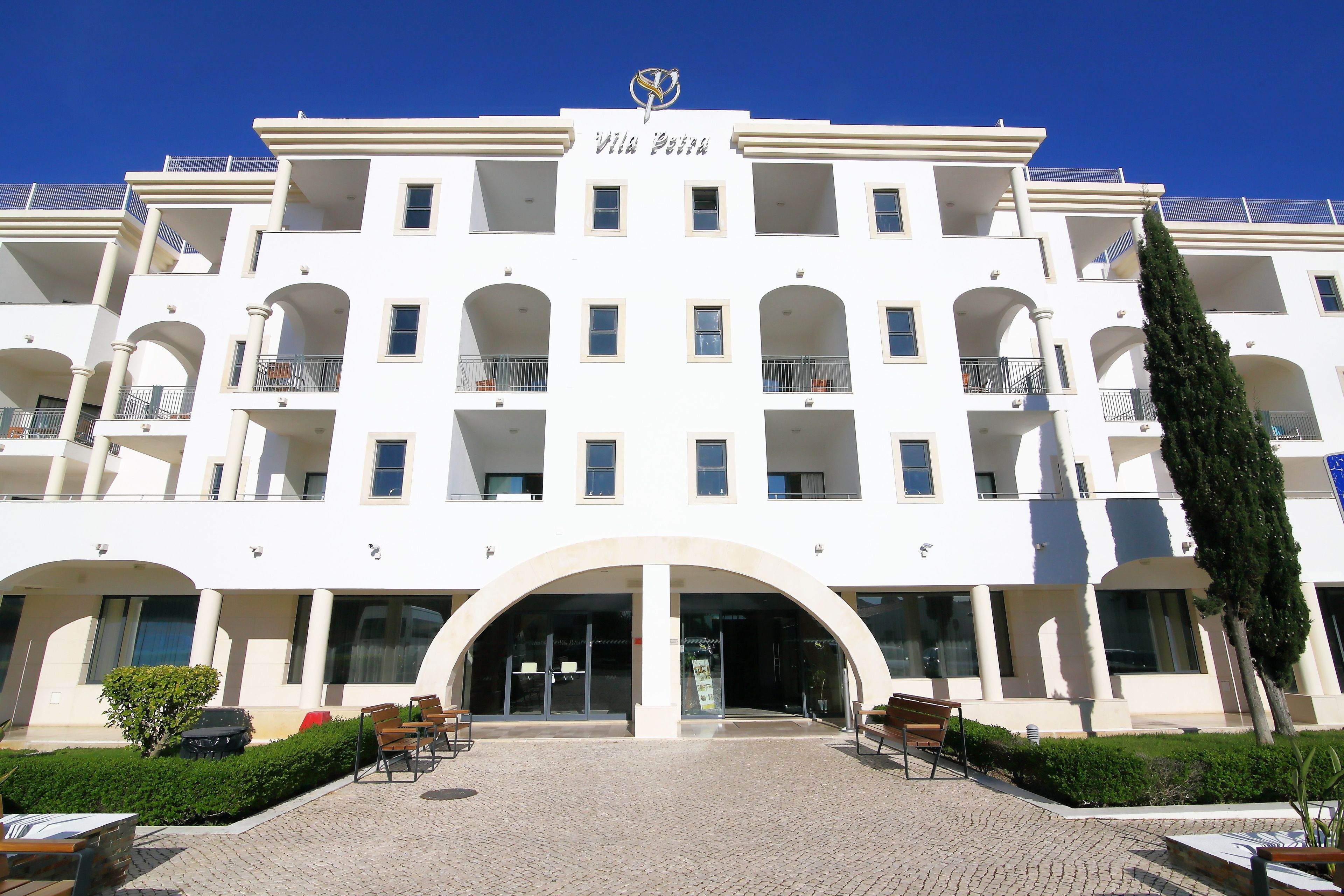 A multi-story hotel with a central entrance, balconies, and a clear blue sky.