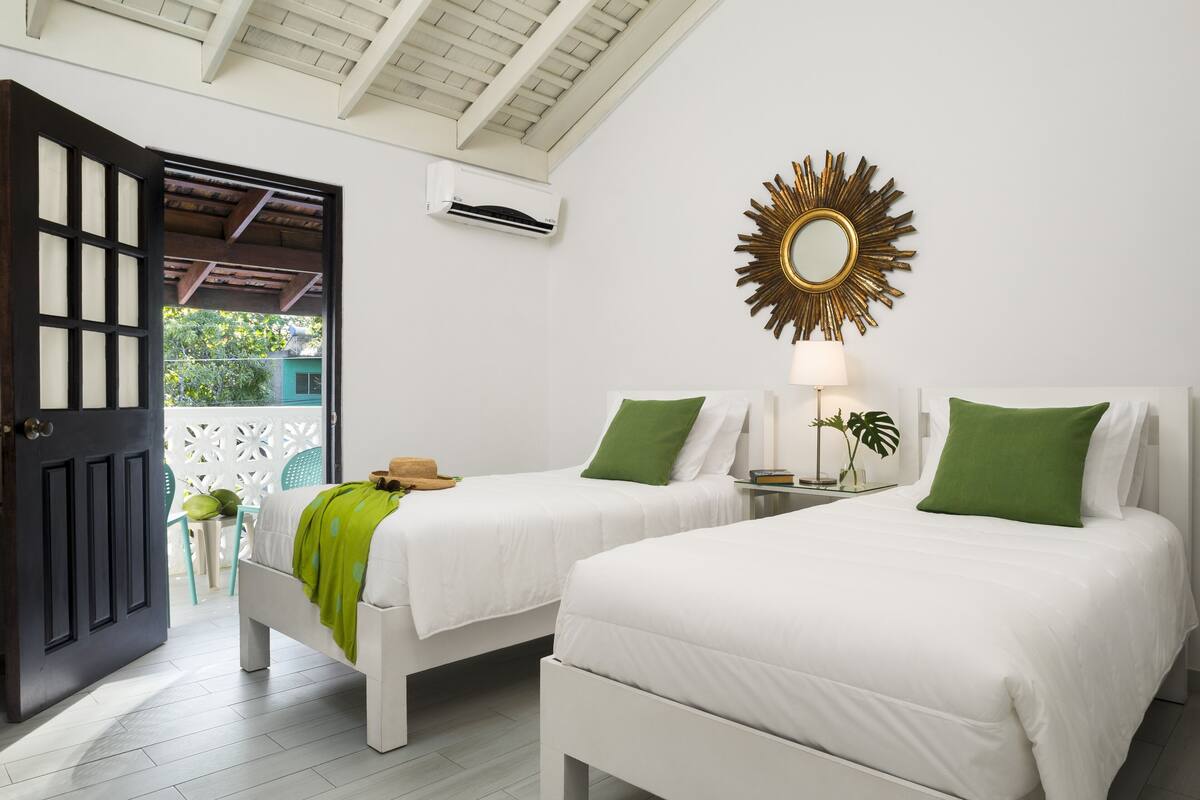 A hotel room with two beds, a ceiling fan, an air conditioning unit, a decorative mirror, and a view of the outside.