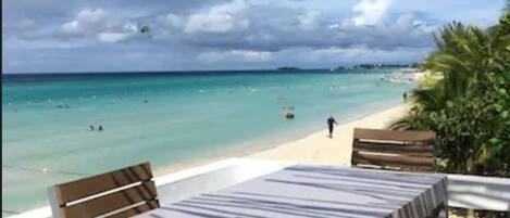 A beachside seating area with a table and chairs, overlooking a turquoise sea and a sandy beach.