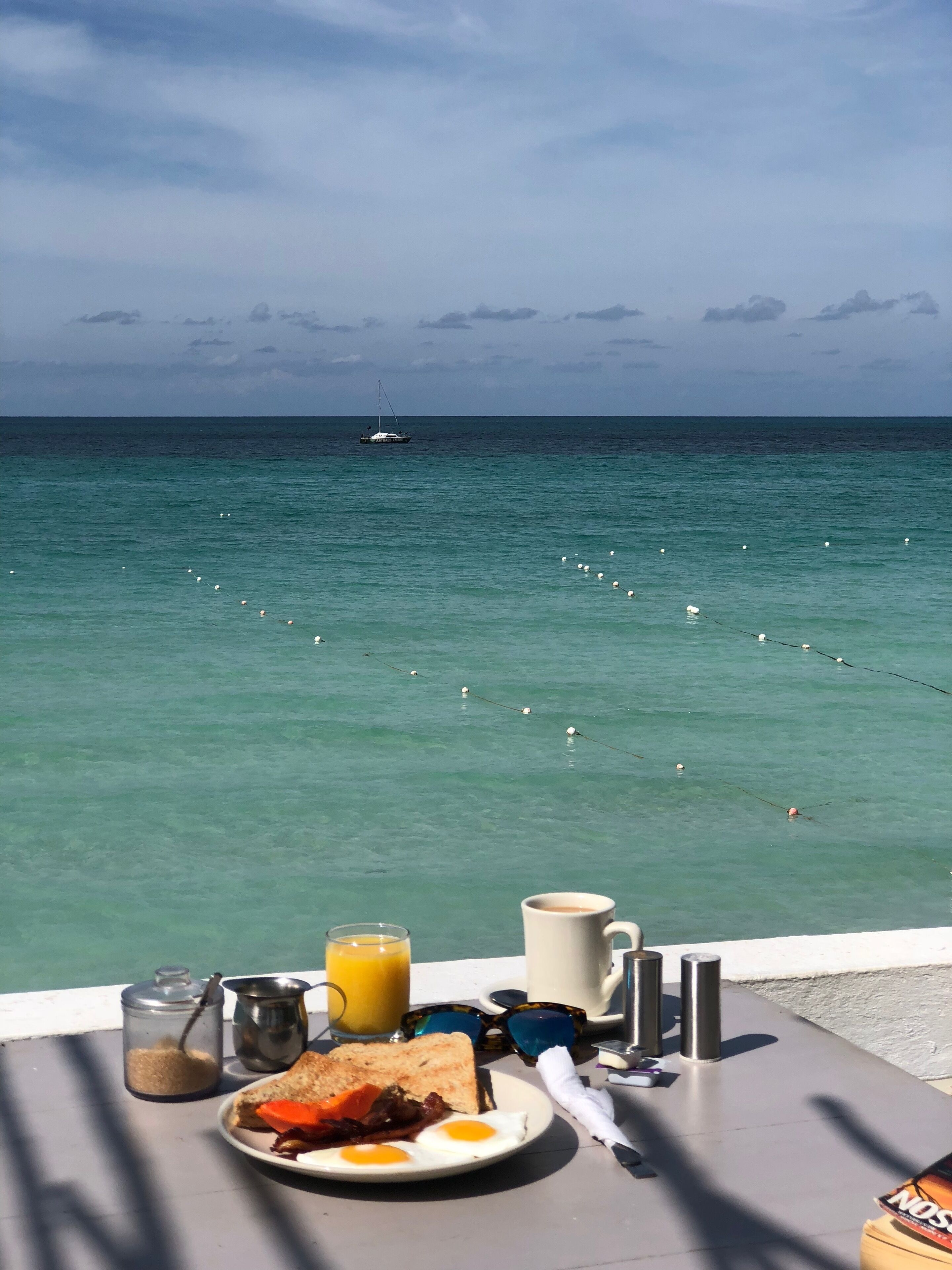 A breakfast table with eggs, toast, and juice, overlooking a calm sea with a boat and birds.