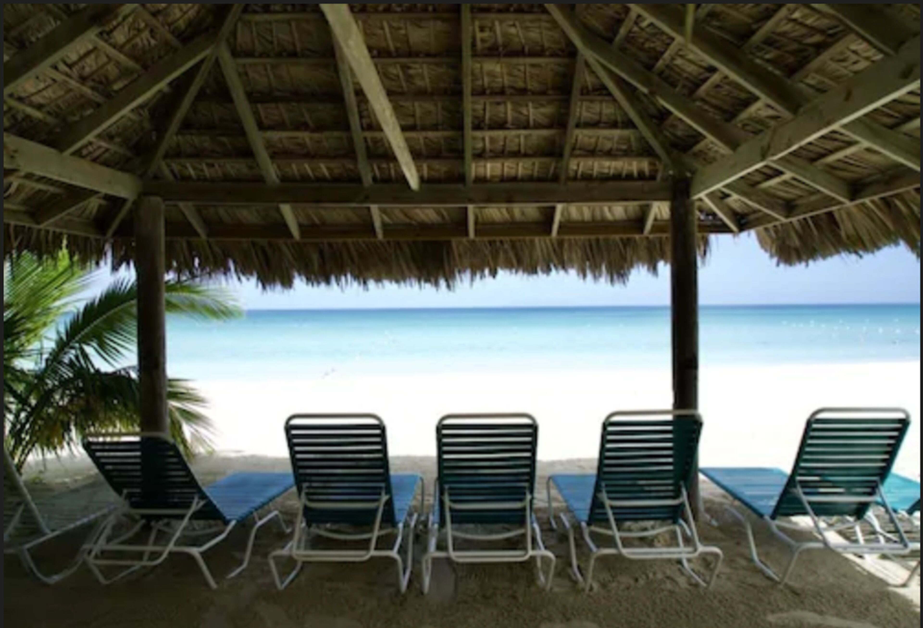 A beach hut with a thatched roof and four lounge chairs facing the ocean.