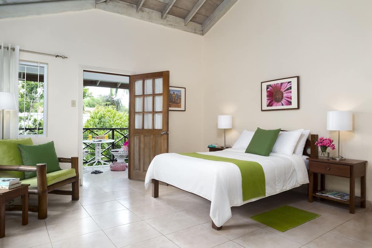A hotel room with a bed, green and white bedding, a wooden nightstand, a green rug, a wooden chair, a small table with a book, and a door leading to an outdoor area with a balcony.