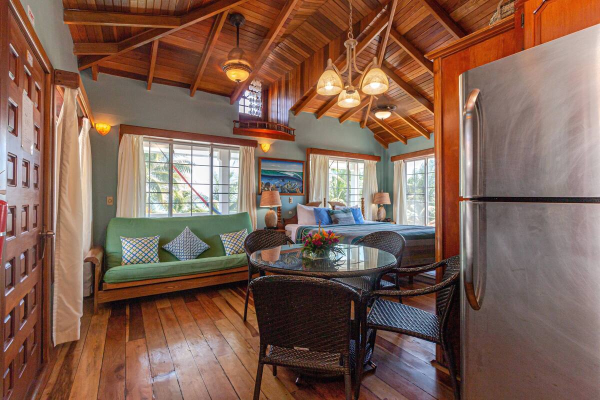 A kitchen with a large stainless steel refrigerator, wooden ceiling beams, a dining table with wicker chairs, and a green sofa with patterned cushions.