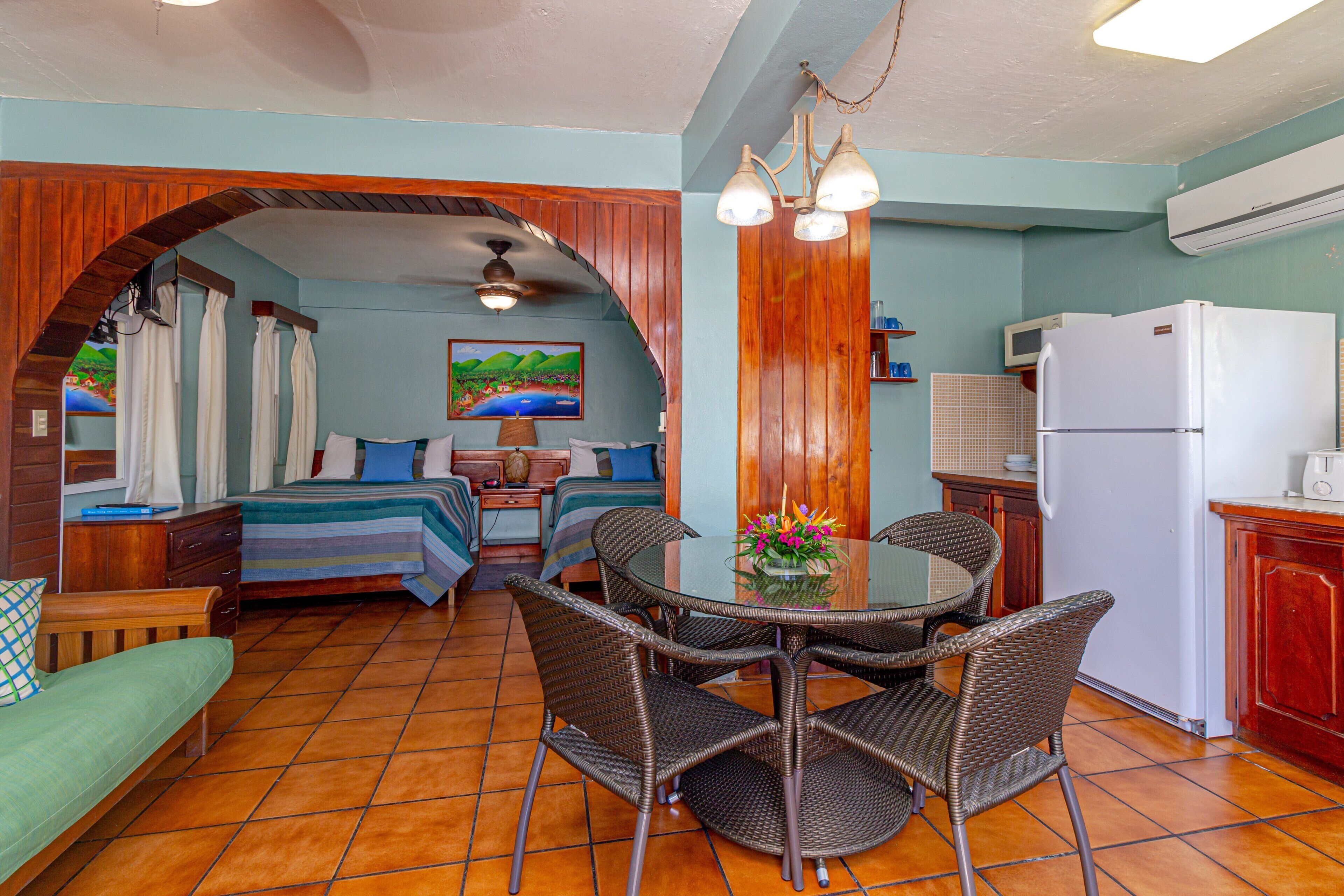 A room with a bed, a sofa, a dining table with chairs, a kitchen area with a refrigerator, and a ceiling fan.