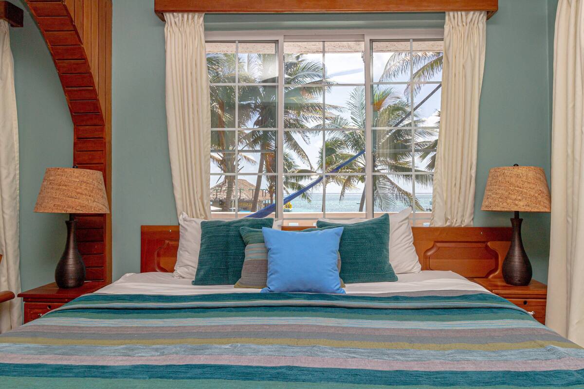 A bed with a striped bedspread, a view of palm trees through a window, and two lamps on either side of the bed.
