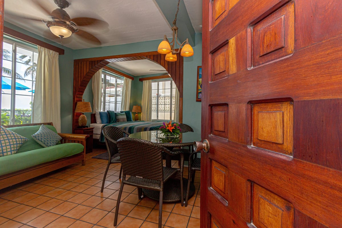 A room with a wooden door, a green sofa, a dining table with chairs, and a ceiling fan.
