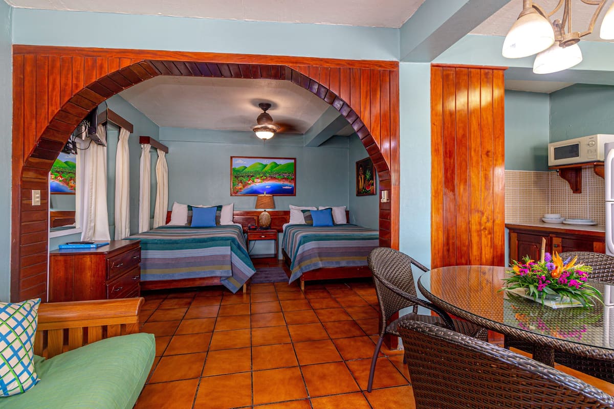 A room with two beds, a dining table with chairs, a ceiling fan, and a small kitchen area.