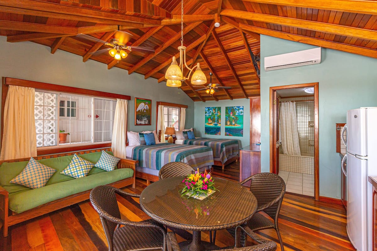 A cozy room with a wooden ceiling, a green sofa with patterned cushions, a round table with wicker chairs, and a bed with striped bedding.