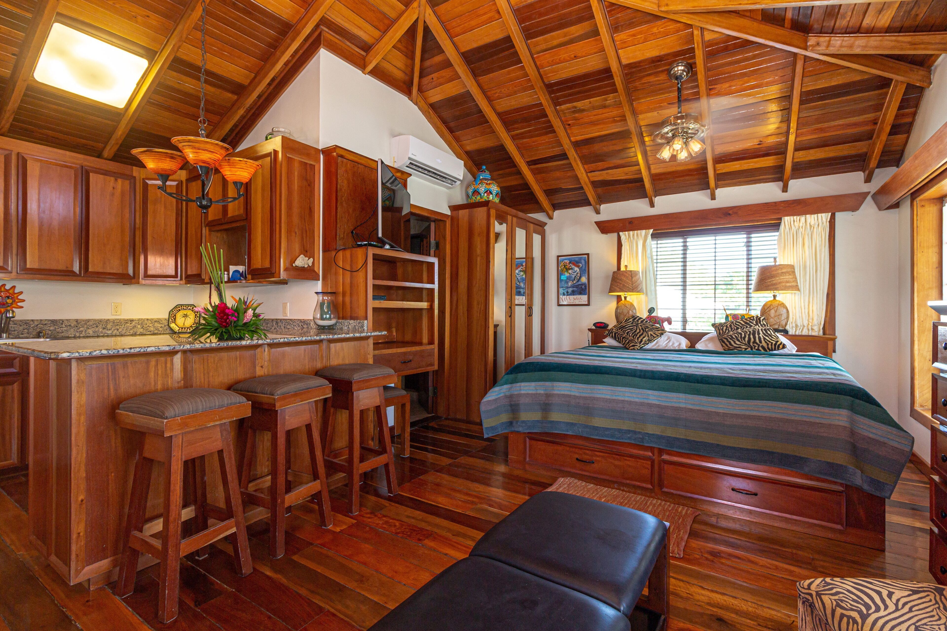 A wooden interior with a kitchen area, bar stools, a bed with a striped bedspread, and a ceiling fan.
