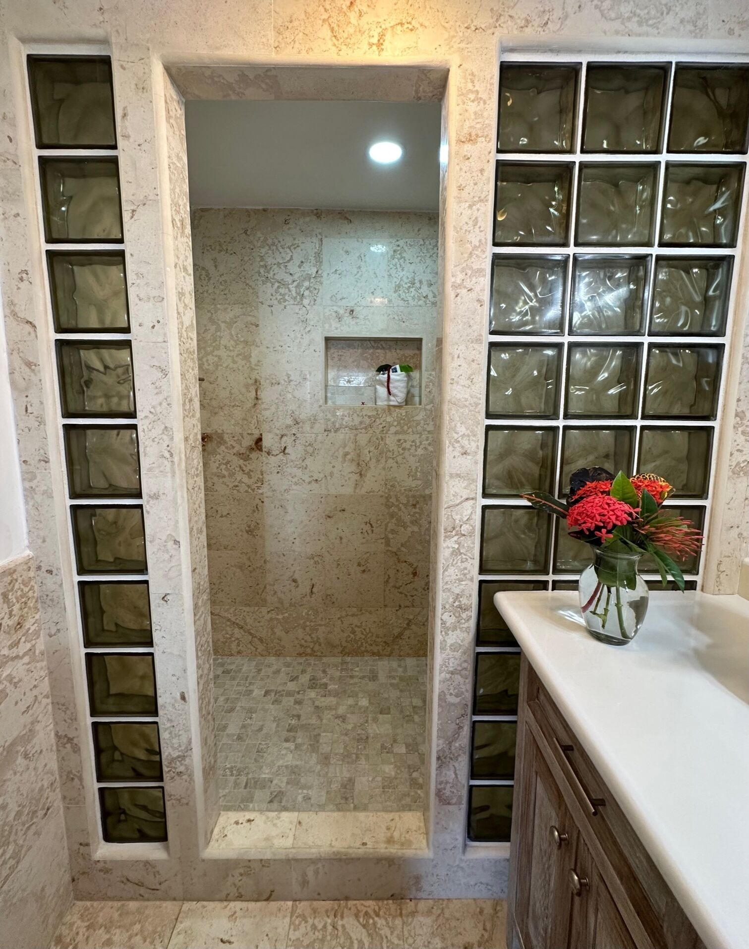 A bathroom with a shower area enclosed by frosted glass panels, a tiled floor, and a wooden cabinet with a vase of flowers on the counter.