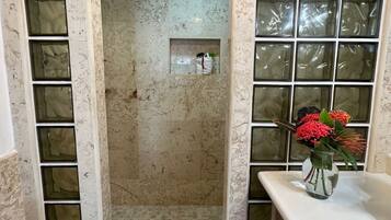A bathroom with a shower area enclosed by frosted glass panels, a tiled floor, and a wooden cabinet with a vase of flowers on the counter.