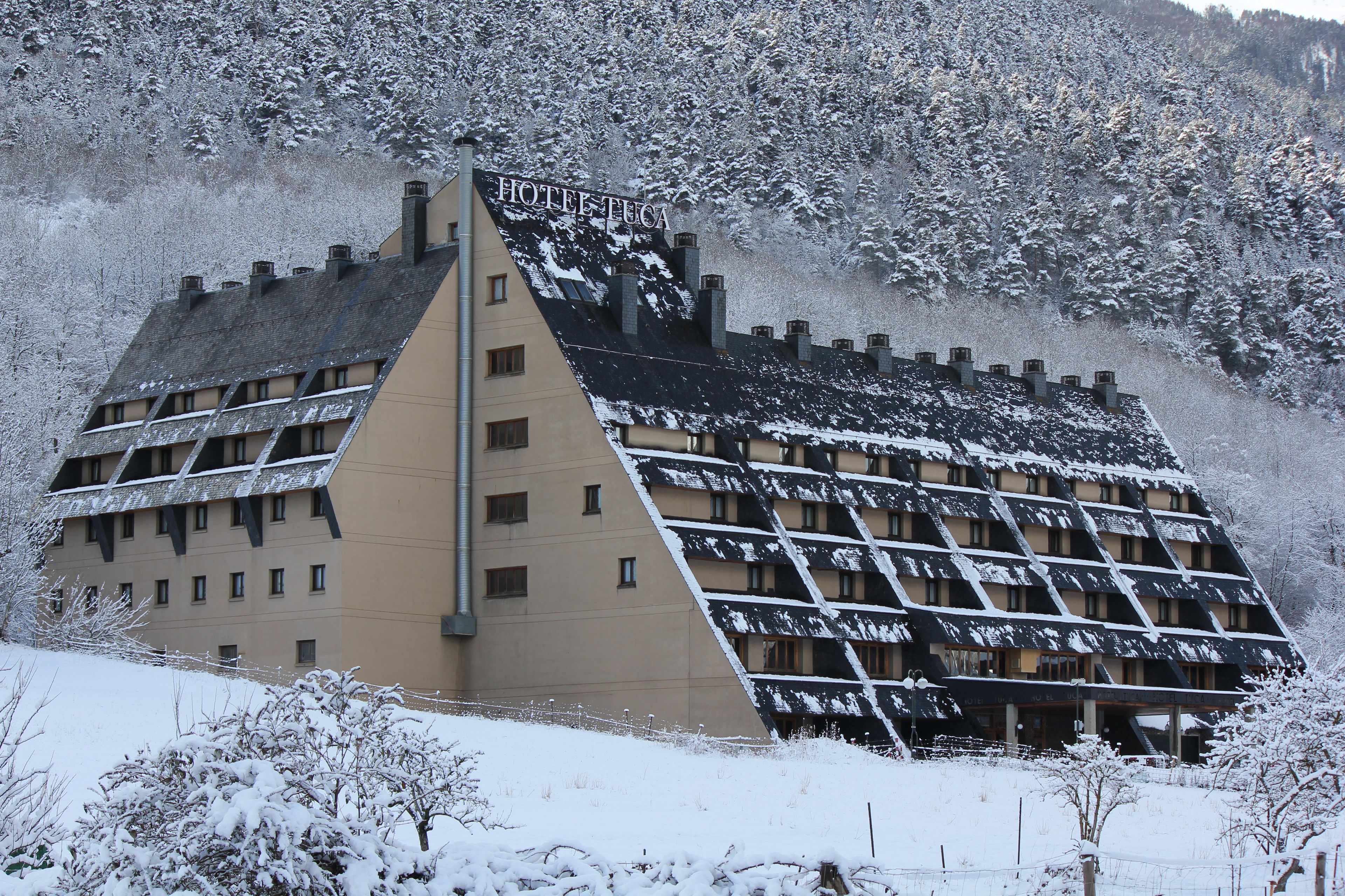 Un hotel con techo inclinado y numerosas ventanas, rodeado de árboles y colinas cubiertas de nieve.