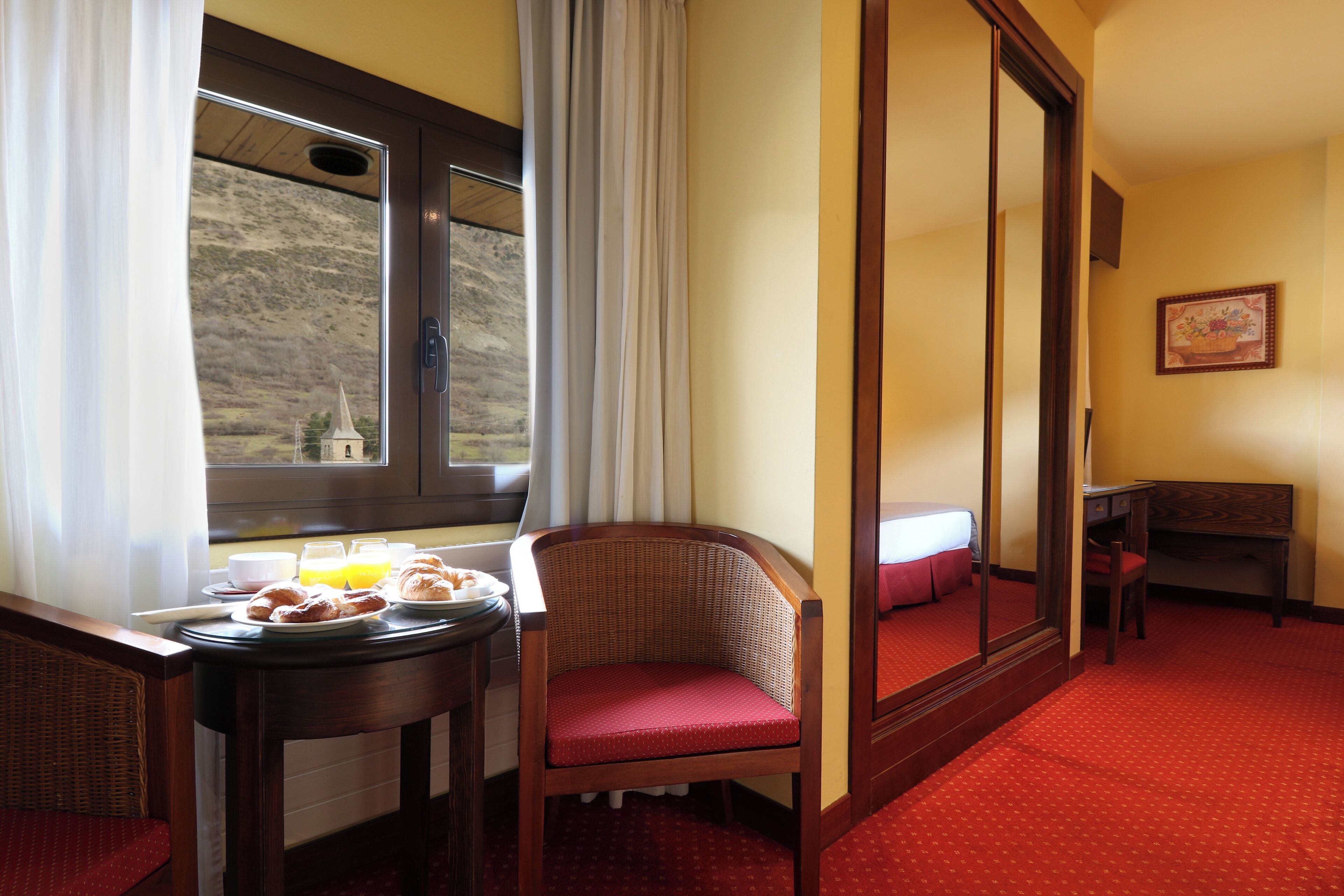Een hotelkamer met een ontbijt gedekte eettafel, een rieten stoel, een spiegel en een klein bureau.