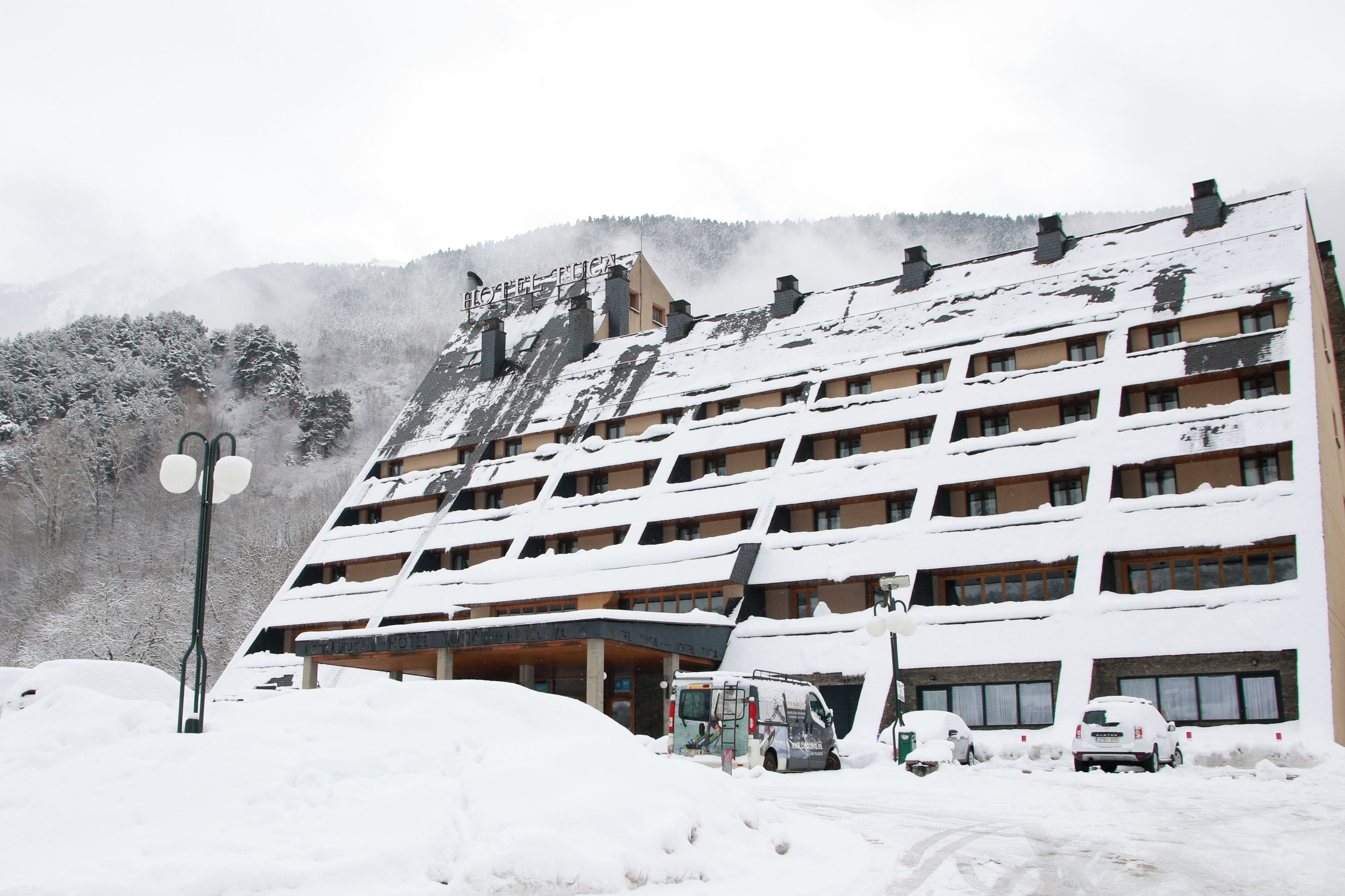 Un hotel de varios pisos con fachada nevada, un estacionamiento con varios automóviles y montañas al fondo.