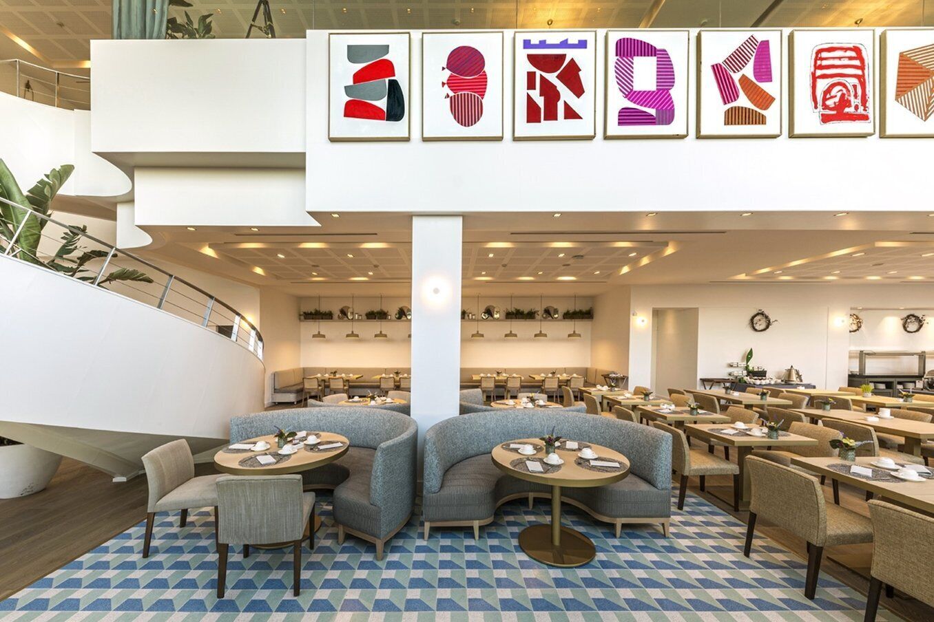 Een moderne eetruimte met ronde tafels en gestoffeerde zitmogelijkheden, een trap en abstracte kunst aan de muur.