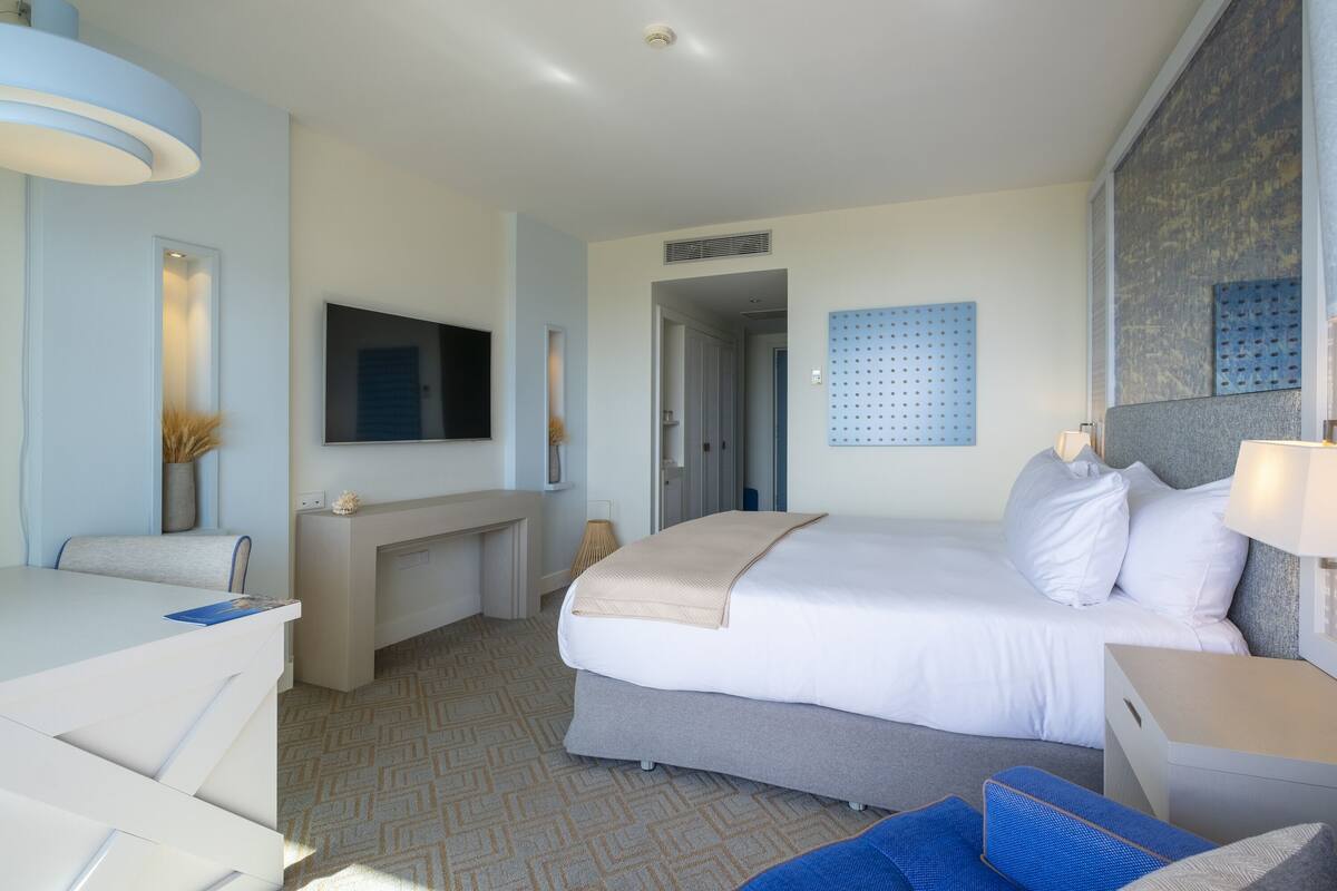 A modern hotel room with a bed, a desk, a chair, a TV, and a large window.
