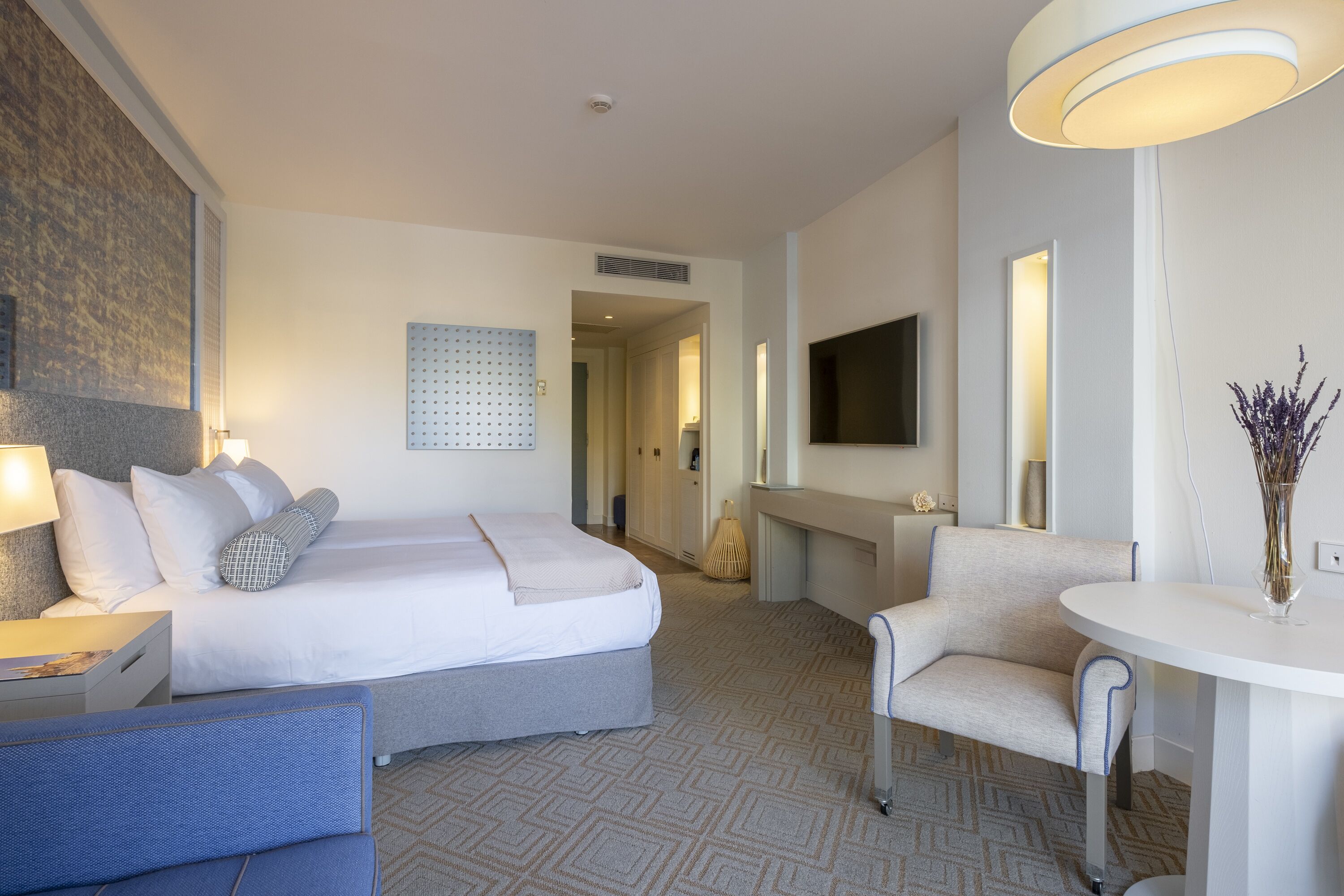 Una habitación de hotel con cama, escritorio, silla, televisor y un florero con flores.
