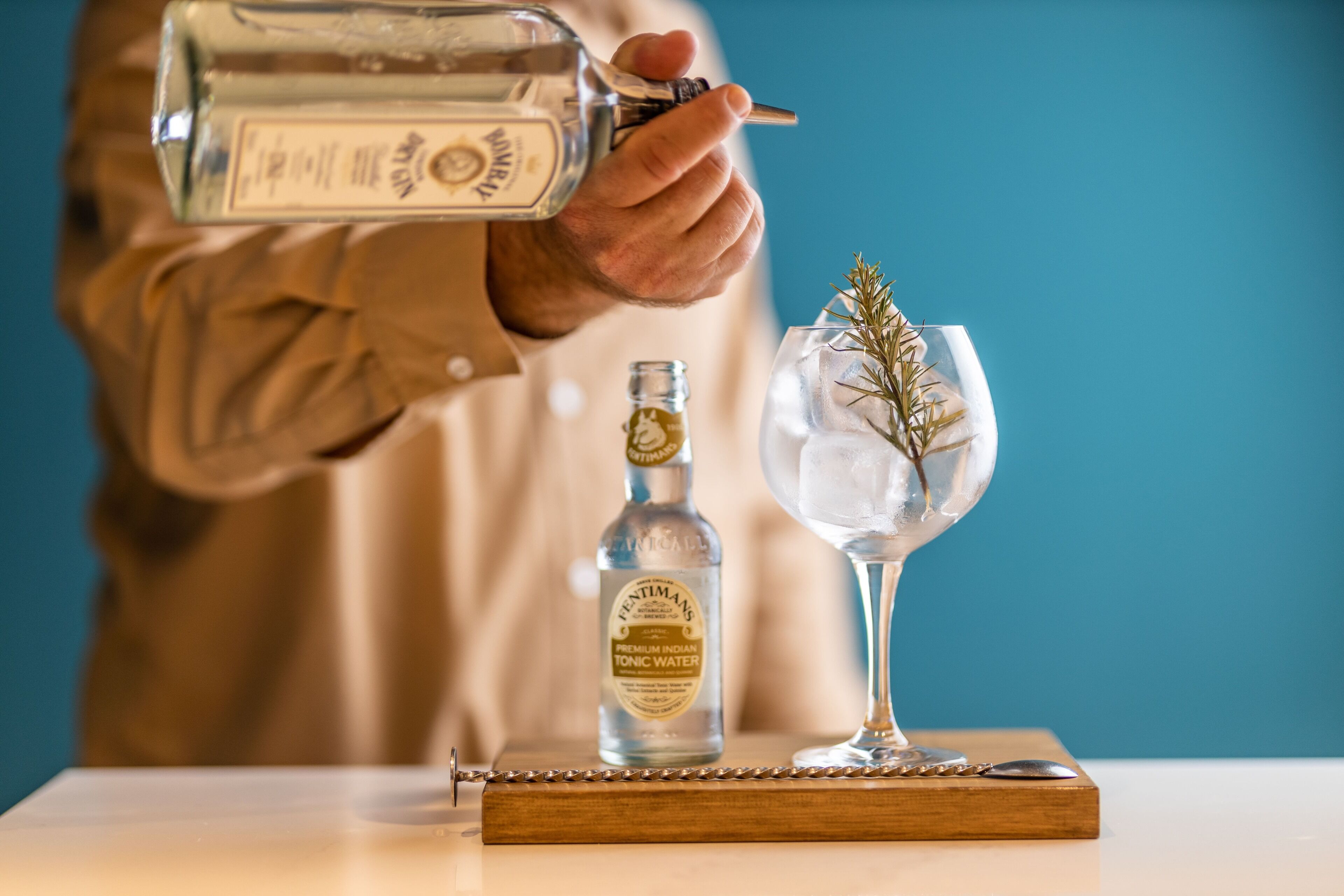 Una persona vertiendo un líquido transparente en un vaso con hielo y una ramita de romero. Se observa una botella de tónica india premium sobre la mesa.