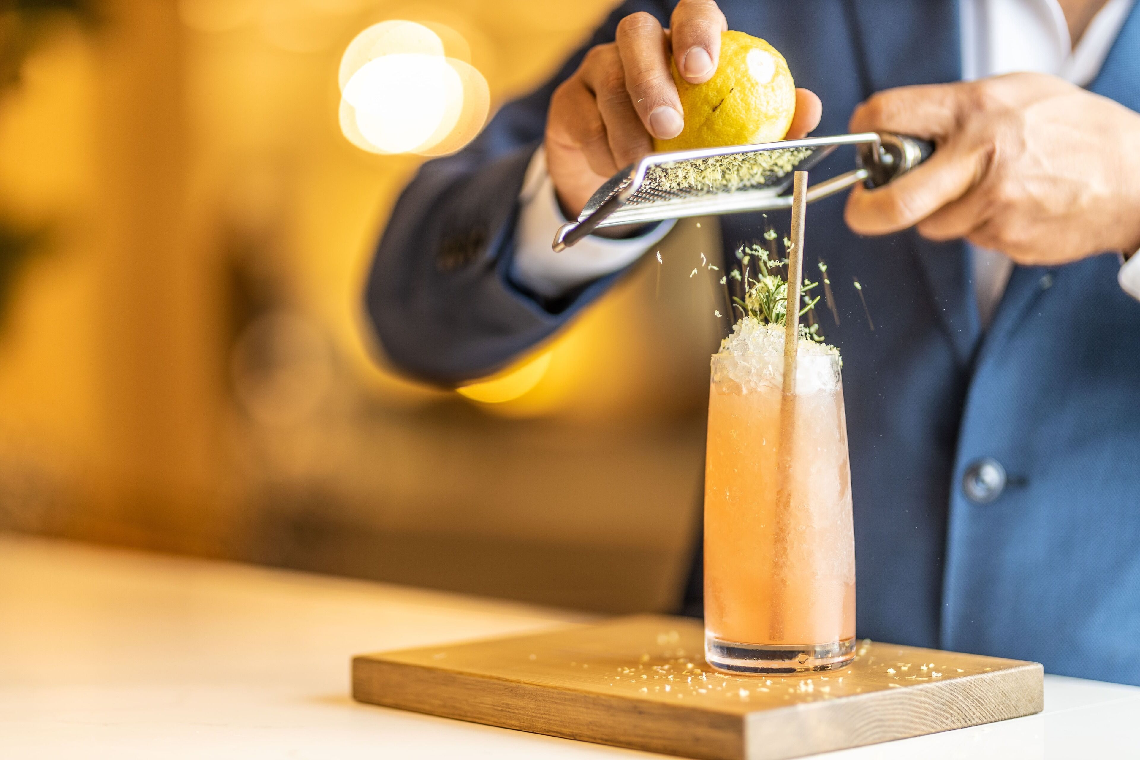Eine Person reibt Zitronenschale über einen Drink in einem hohen Glas.