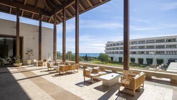 Un espacio de estar techado al aire libre con mobiliario moderno, ventanas amplias y vistas a un edificio y paisaje.