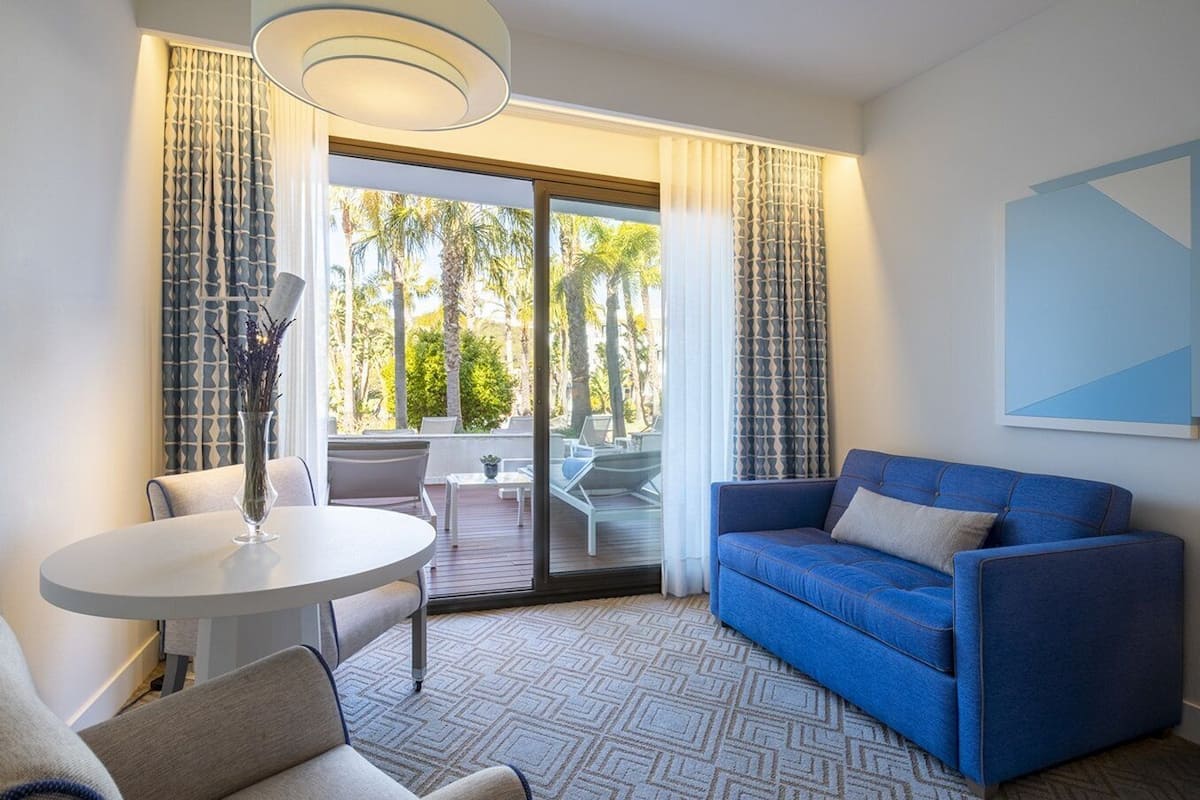 A modern hotel room with a blue sofa, a round table, and a balcony with a view of palm trees.