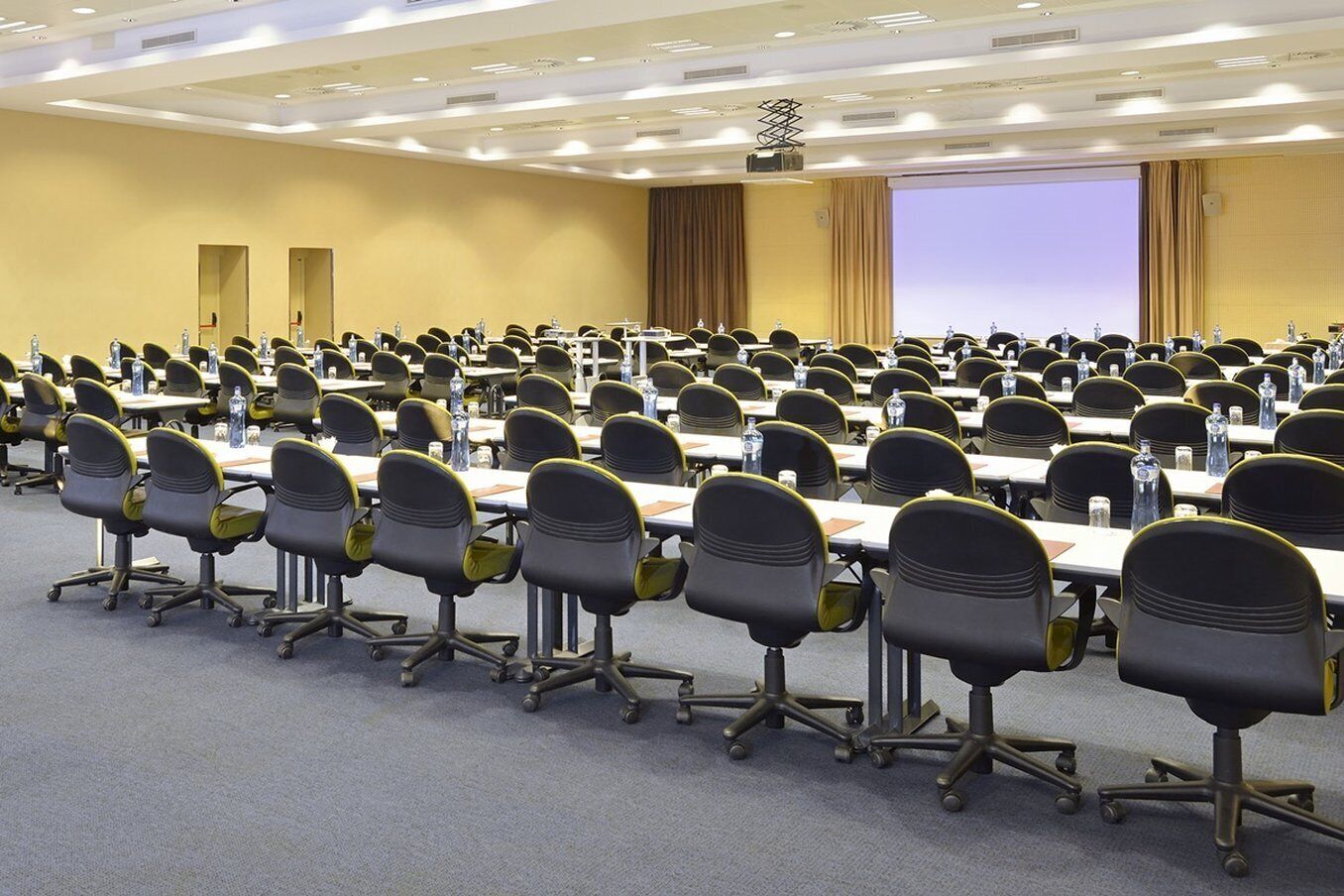 Una vasta sala conferenze con file di tavoli e sedie, dotata di bottiglie d'acqua e cartellini.