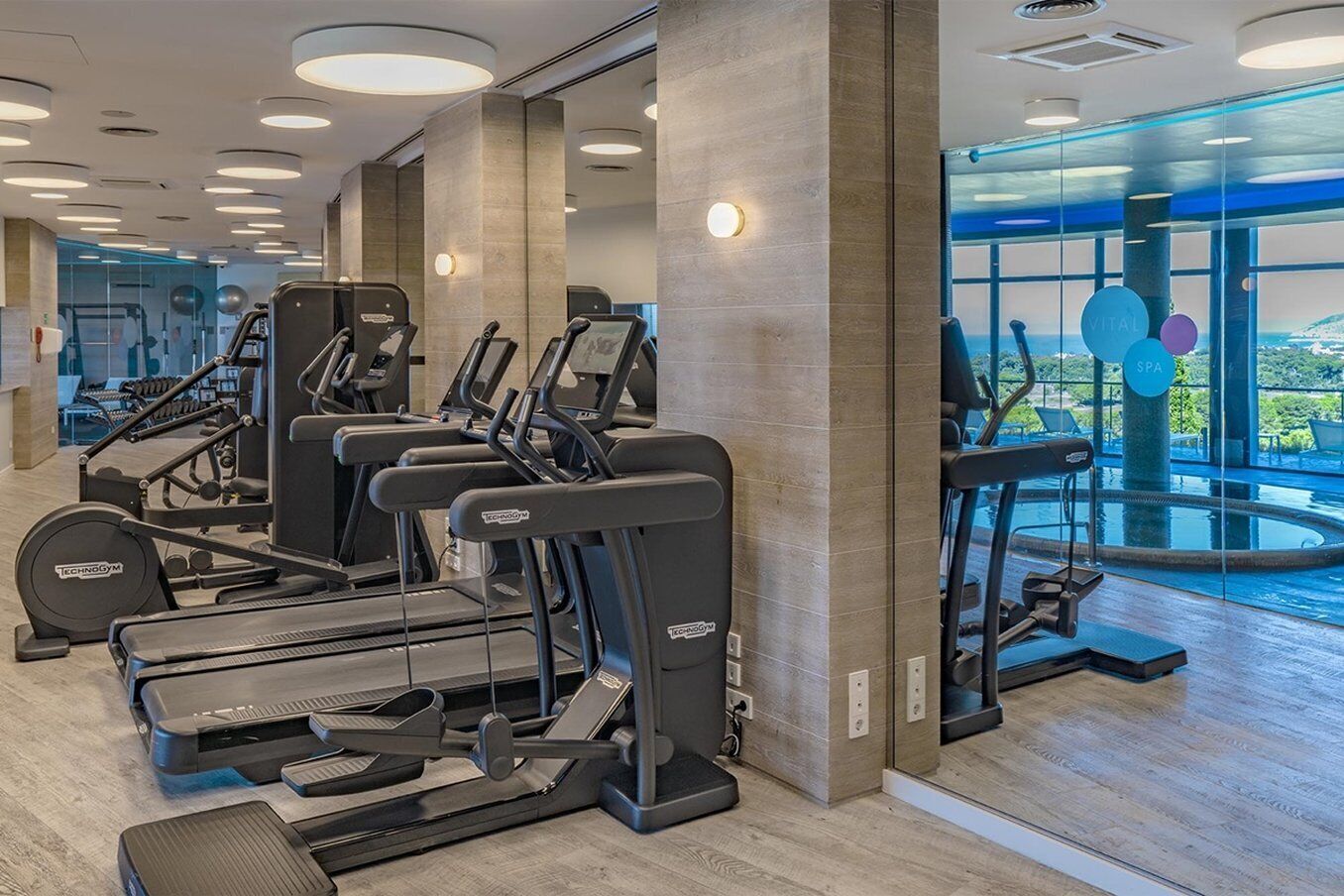 A modern gym with various cardio machines, a mirror wall, and a view of a pool area.