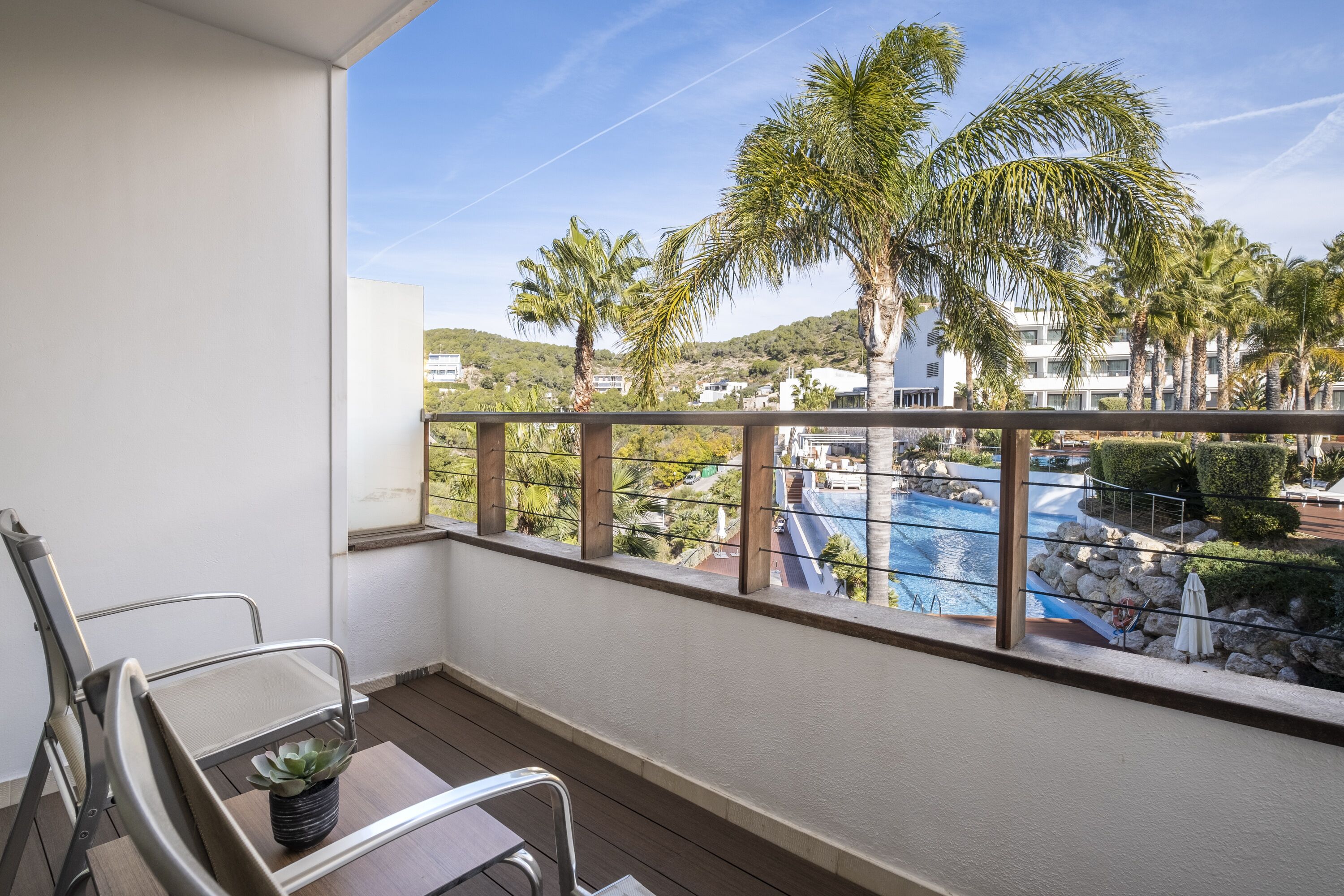 A balcony with a view of a pool, palm trees, and a hilly landscape.