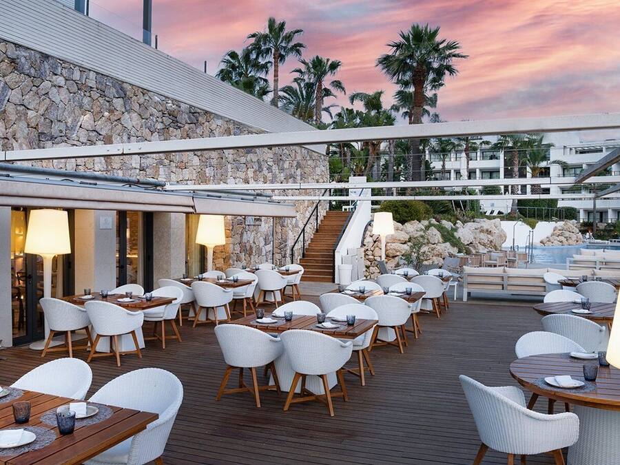 Un espace de restauration aménagé sur le toit, avec des chaises et des tables en osier blanc, un mur en pierre et une vue sur des palmiers et une piscine.