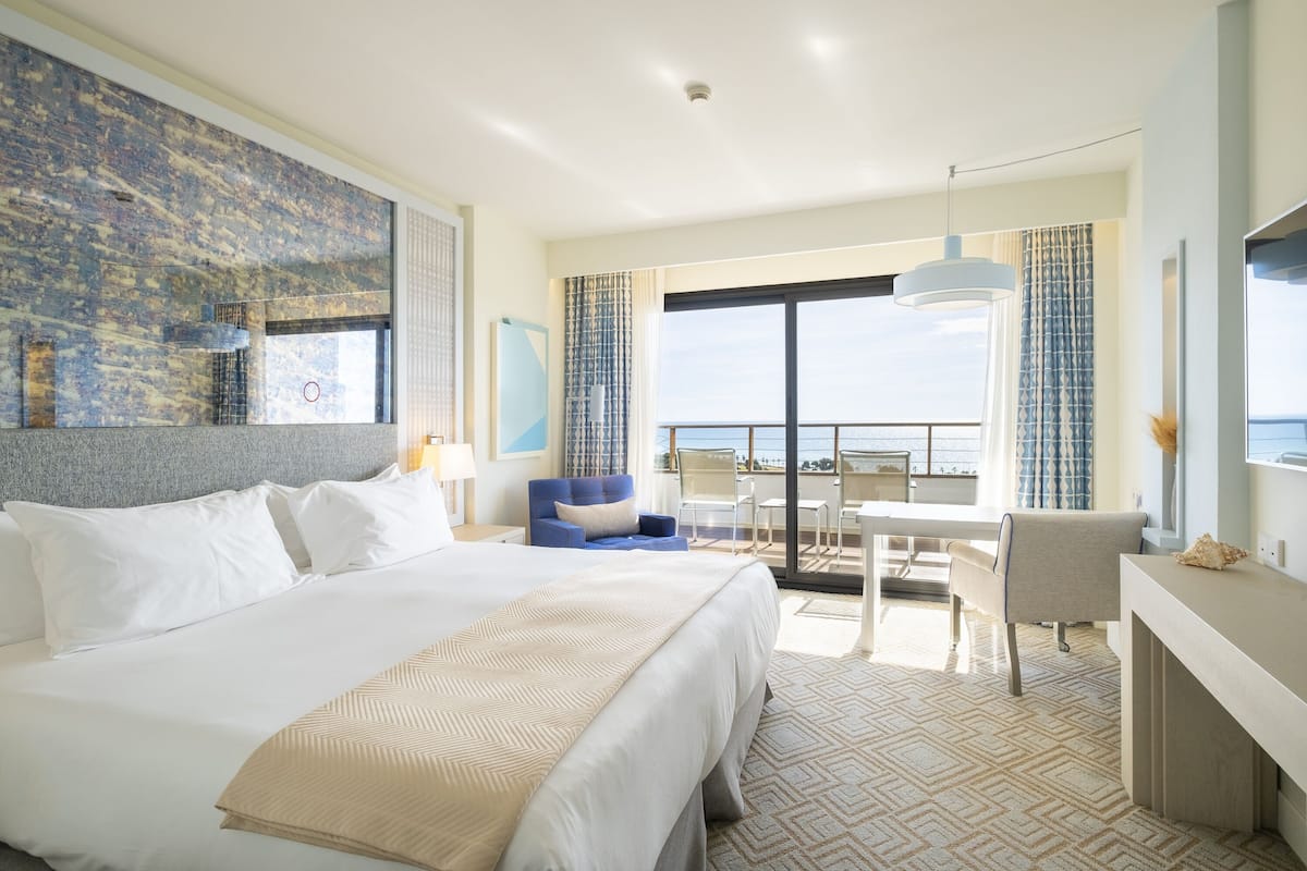 A hotel room with a large bed, a desk, a chair, and a balcony with a view of the sea.