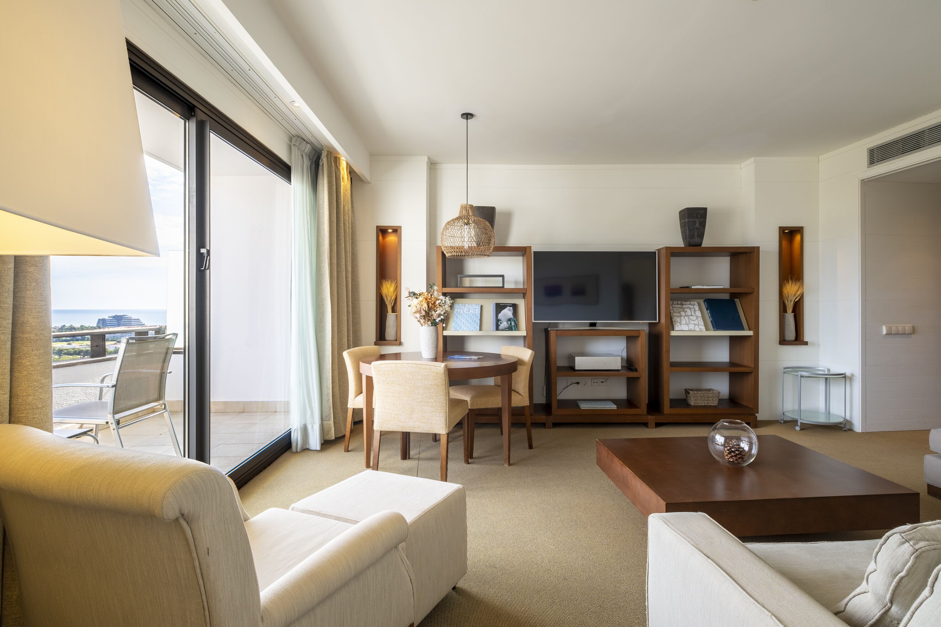 A modern hotel room with a balcony, a dining area, and a living room with a TV, bookshelves, and a coffee table.