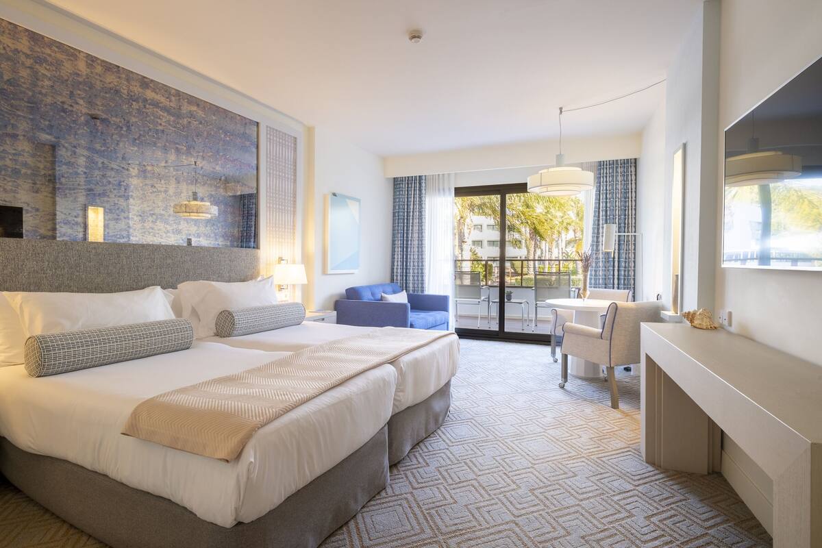 A hotel room with a large bed, a desk, a chair, and a balcony with a view of buildings and trees.