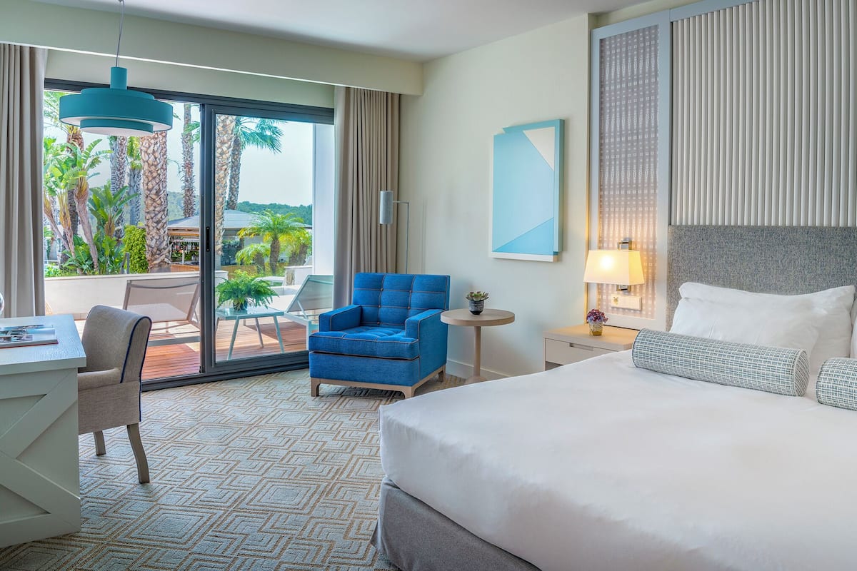 A hotel room with a bed, a desk, a chair, a small table, and a view of palm trees through the glass door.