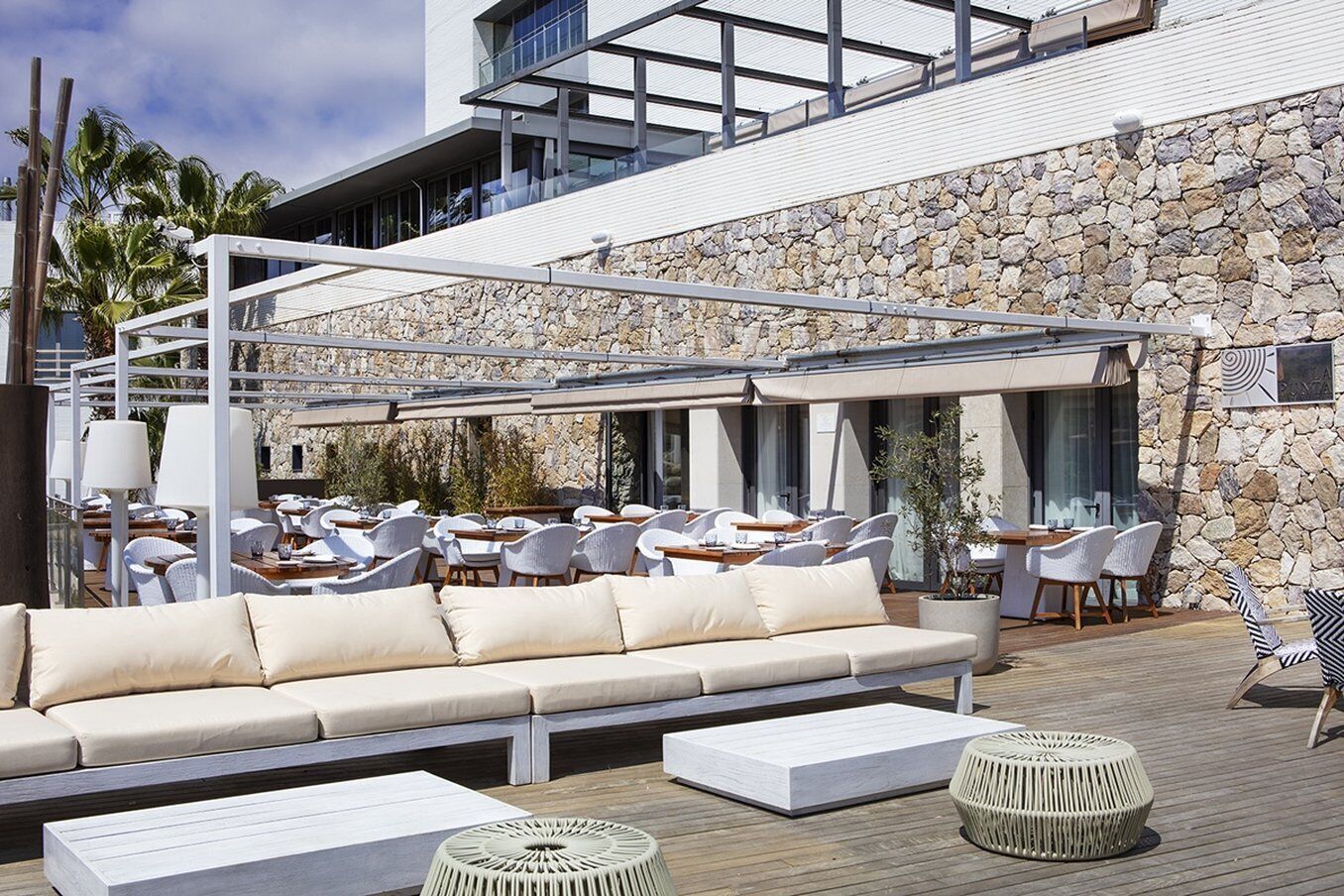 Un espace extérieur aménagé avec un canapé, des fauteuils et des tables, offrant une vue sur un mur en pierre et un bâtiment moderne.