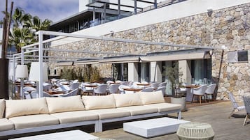 An outdoor seating area with a sofa, chairs, and tables, overlooking a stone wall and a modern building.