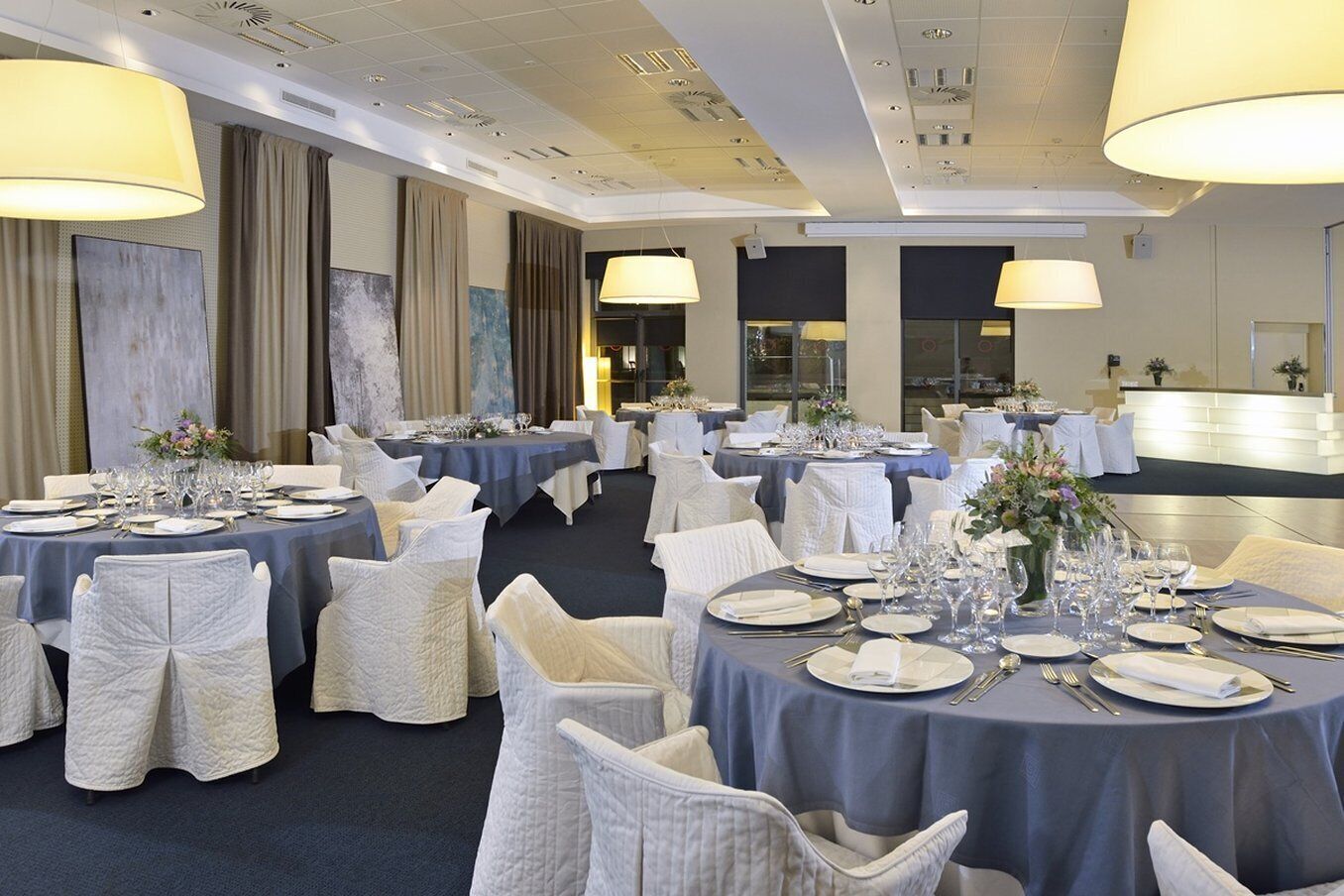 Un salón de banquetes con mesas redondas dispuestas para comer, sillas blancas y lámparas grandes.