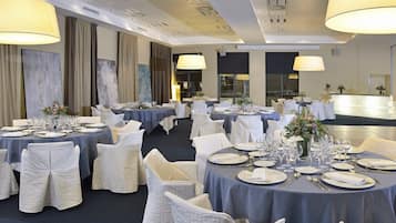 Un salón de banquetes con mesas redondas dispuestas para comer, sillas blancas y lámparas grandes.