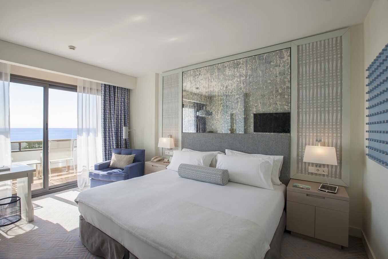 A hotel room with a large bed, a desk, and a chair. There is a balcony with a view of the ocean.
