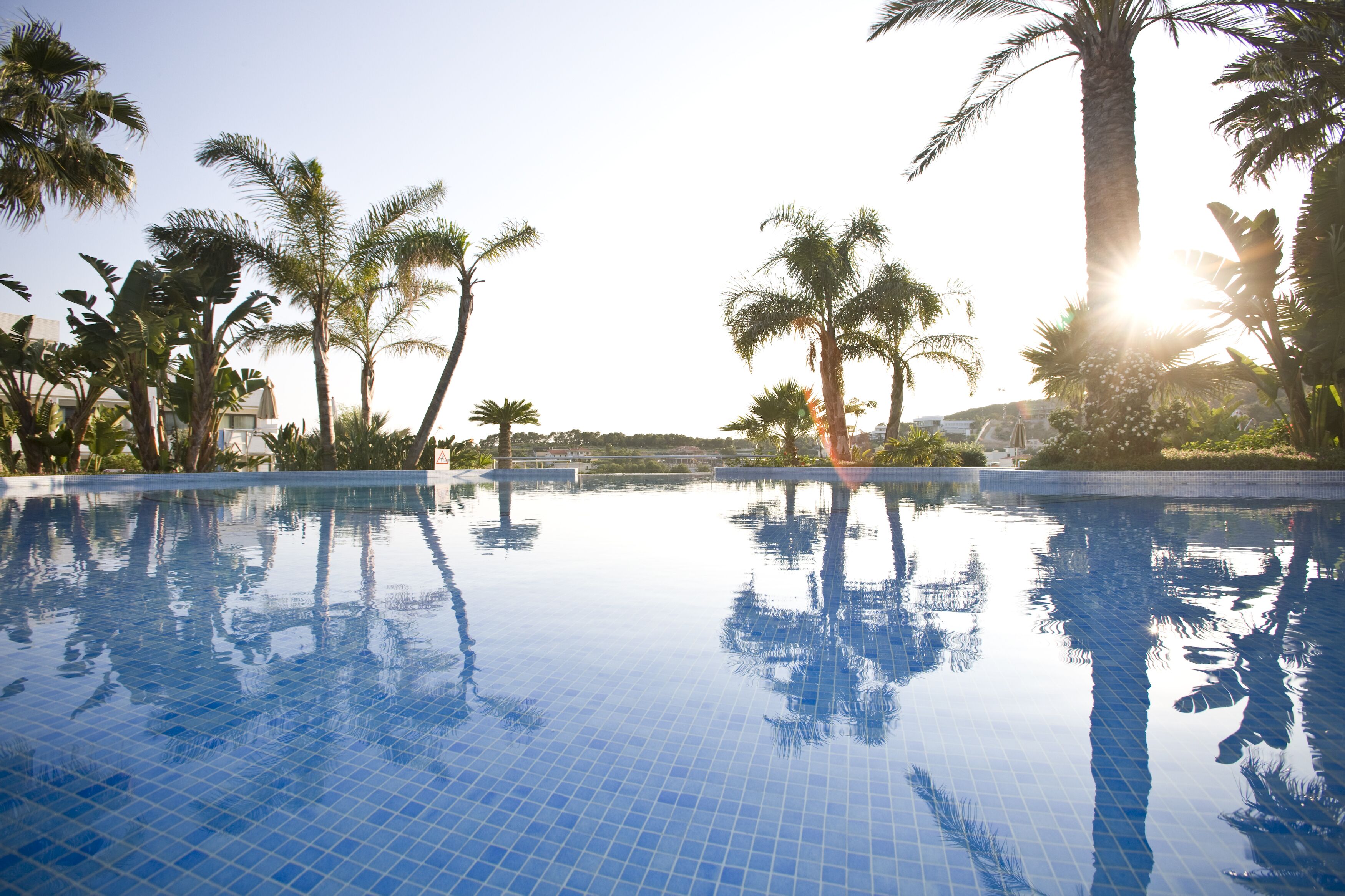 Une piscine d’un bleu éclatant, entourée de palmiers, avec un soleil radieux en arrière-plan.