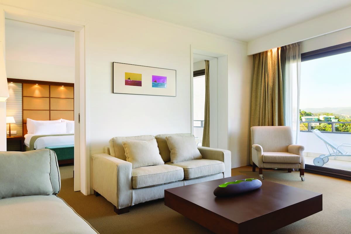 A modern hotel room with a sofa, armchair, and a bed with a view of the city.