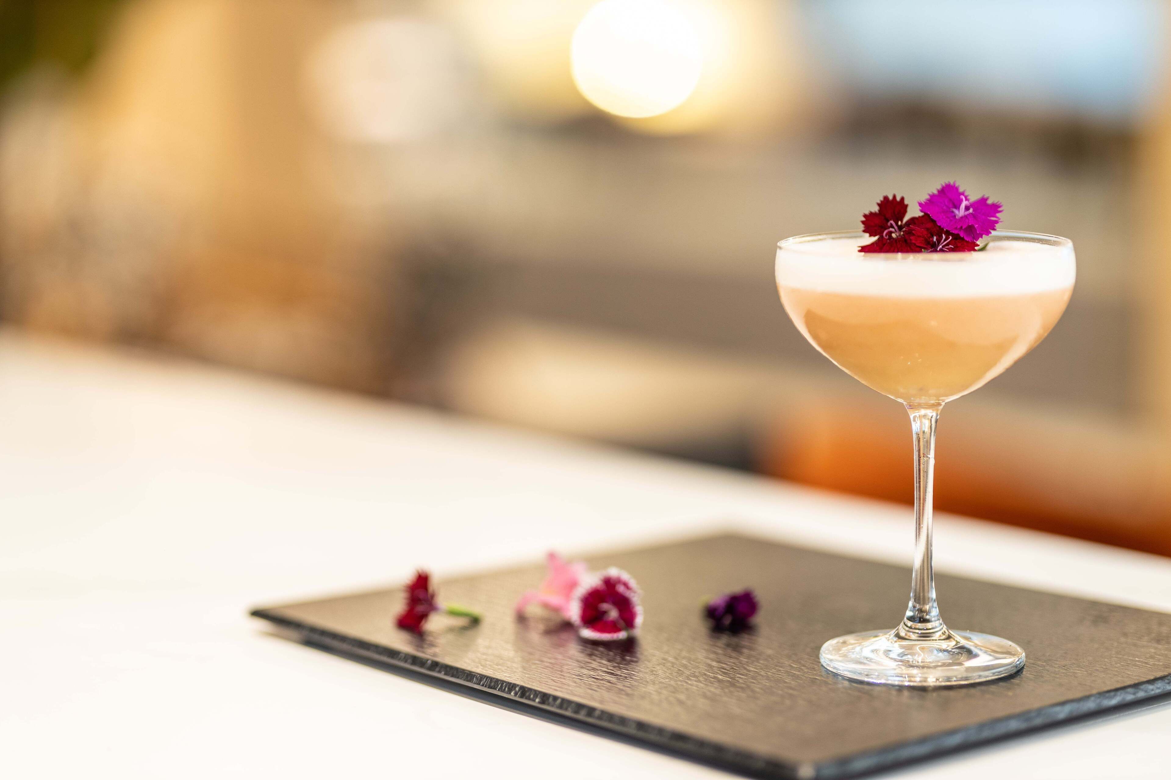 Un cóctel en una copa de martini con espuma, sobre un posavasos de pizarra con pétalos de flores esparcidos.