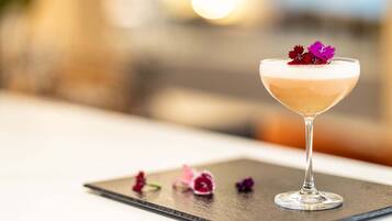 Un cóctel en una copa de martini con espuma, sobre un posavasos de pizarra con pétalos de flores esparcidos.