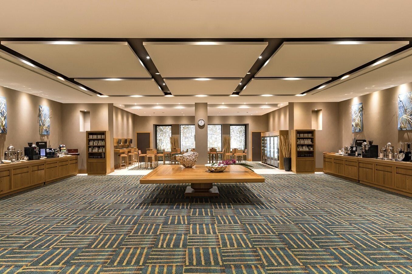 A spacious hotel lobby with a central wooden table, seating area, and a patterned carpet.