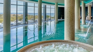 An indoor swimming pool with clear views of the landscape, large pillars, and a hot tub area.