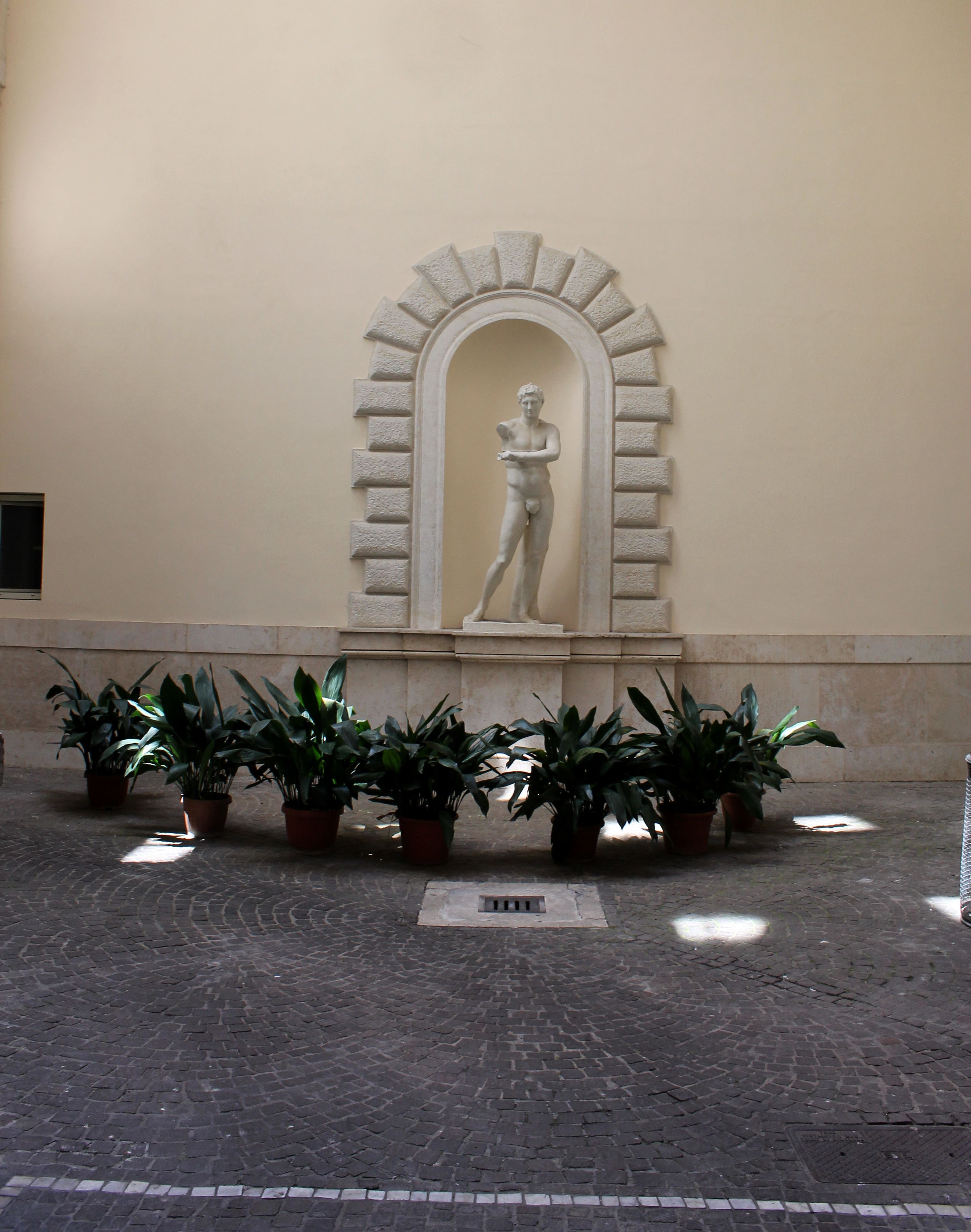 Un patio con una estatua clásica en un nicho abovedado, rodeado de plantas en macetas.
