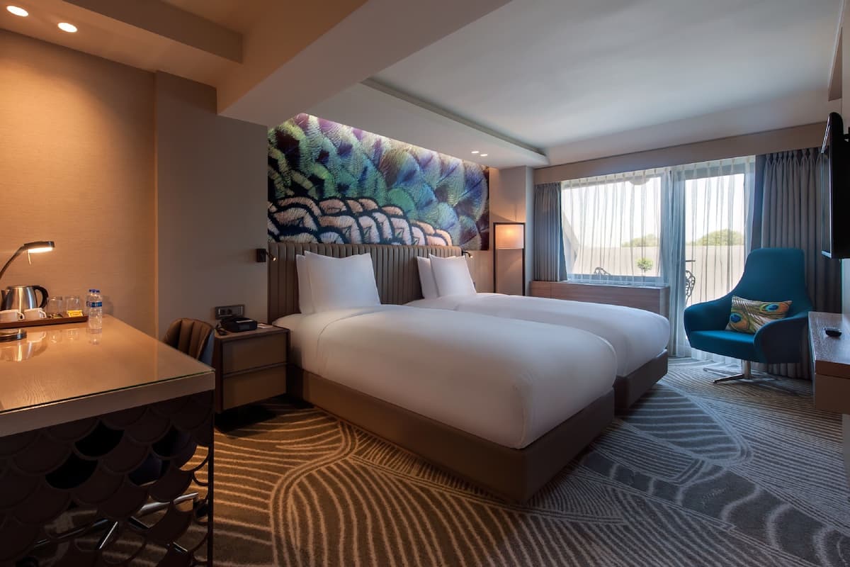 A hotel room with a large bed, a desk with a lamp, a chair, and a large window with curtains.