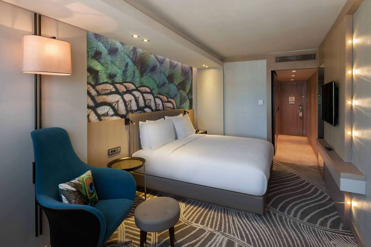 A modern hotel room with a large bed, a blue armchair, a small round table, and a wall-mounted TV.