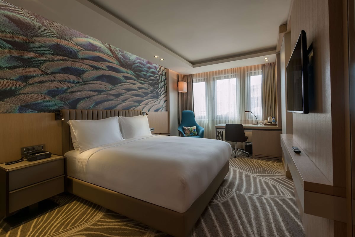 A hotel room with a large bed, a desk with a chair, a television, and a large window with curtains.
