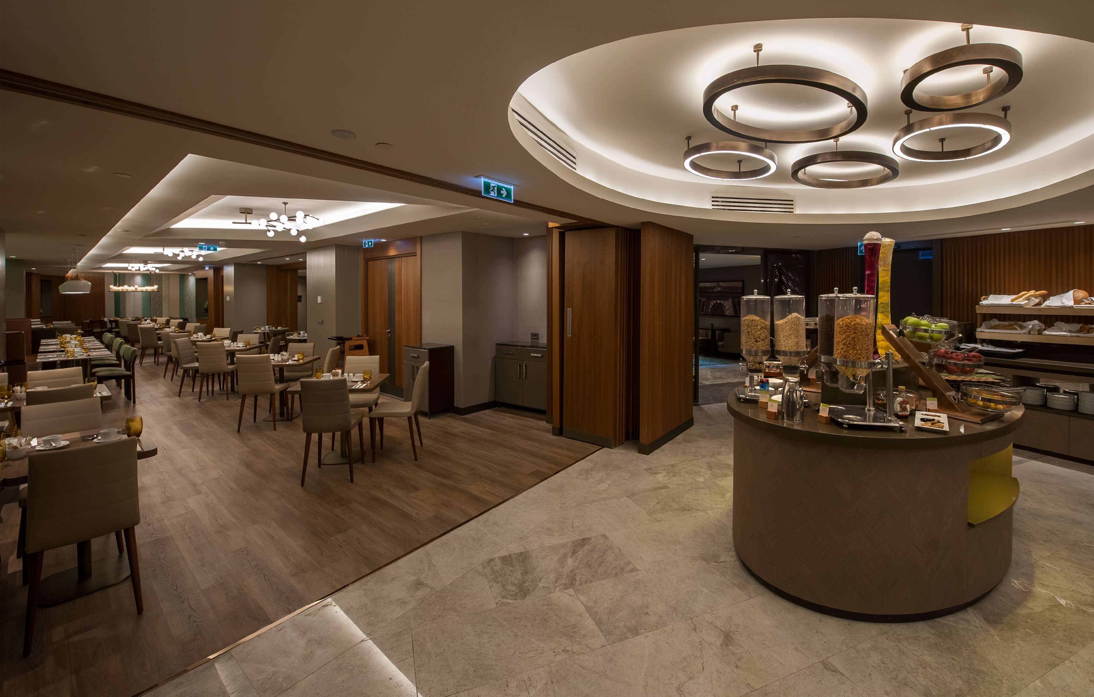 A hotel dining area with multiple tables and chairs, a buffet counter with food items, and modern lighting fixtures.