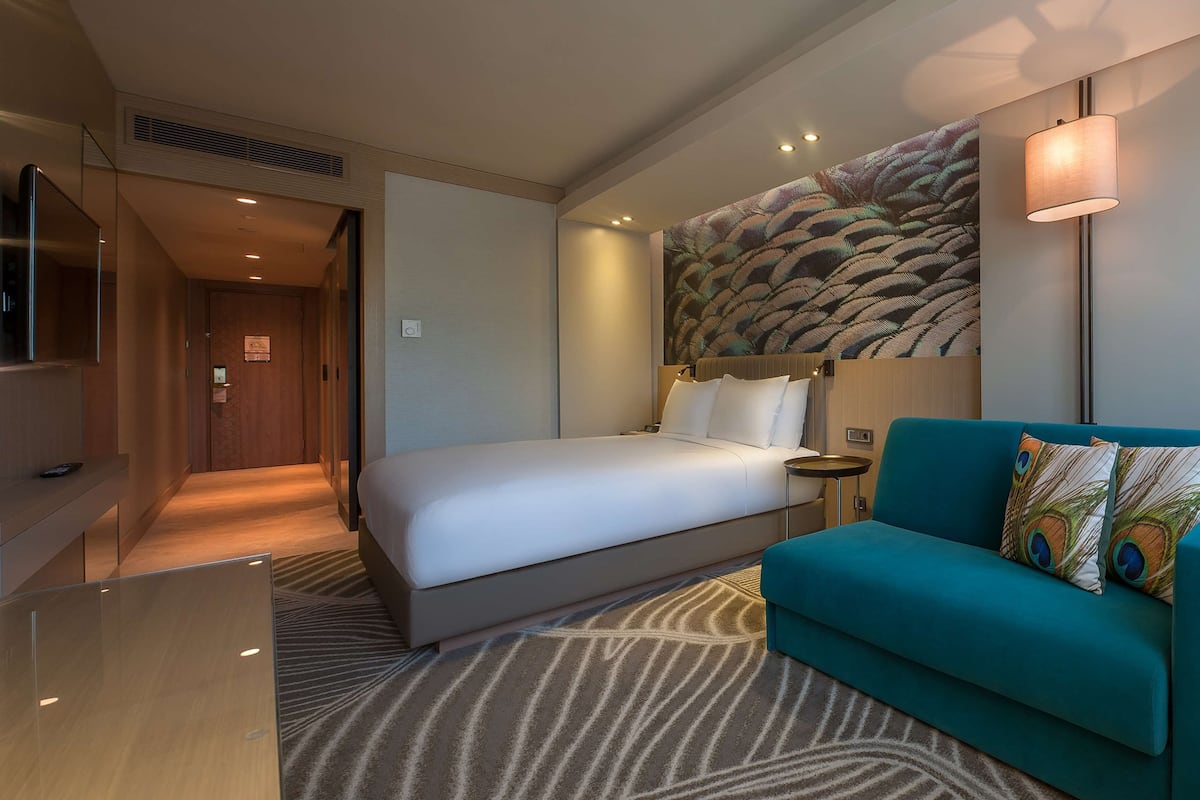 A hotel room with a bed, a teal sofa, a wall-mounted painting, a lamp, and a television.